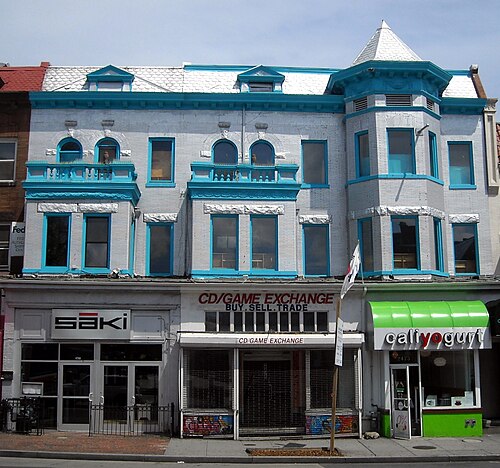 A pale blue Victorian shopfront row on Eighteenth Street, printed shop signs and a green awning over the ground floor units, a roll down security gate pulled partway across one storefront.