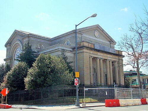 A limestone former church with a full width Corinthian portico and pediment, construction fencing and barriers along the sidewalk in front.