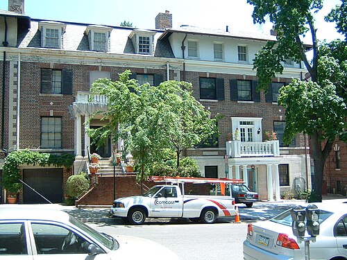 A Mediterranean Revival brick house on Nineteenth Street, dormer windows and a stone balcony over the entrance, a utility truck parked at the curb.