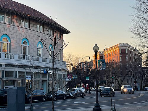 A limestone corner building with a curved second floor balcony at dusk, a brick apartment building glowing pink in the sunset behind it.