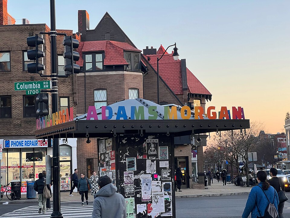 The corner of Eighteenth Street and Columbia Road NW under the neighborhood's own lettered sign, a Victorian building with a red tile roof behind it and pedestrians crossing at dusk.