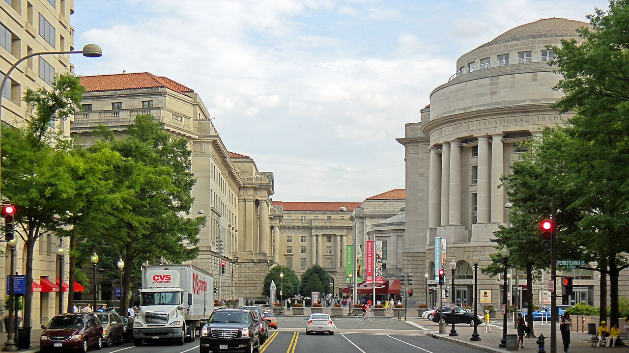 A wide street view down Pennsylvania Avenue NW, limestone neoclassical buildings on both sides with the Ronald Reagan Building's columned rotunda on the right.