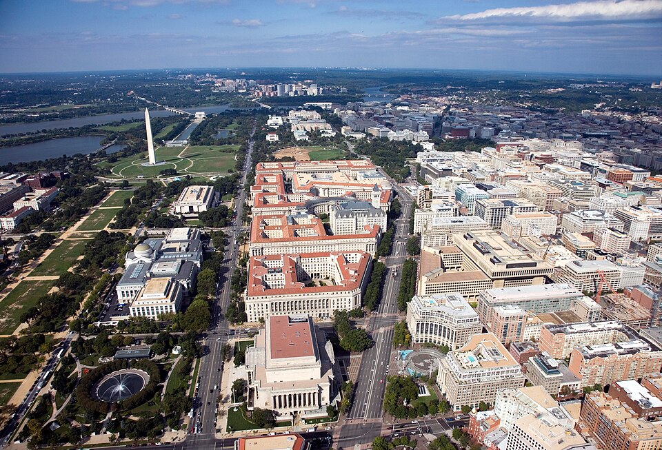 An aerial view of the Federal Triangle wedge, the National Archives Building's domed colonnaded entrance visible mid frame with red tile roofs beyond it and the Washington Monument in the distance.