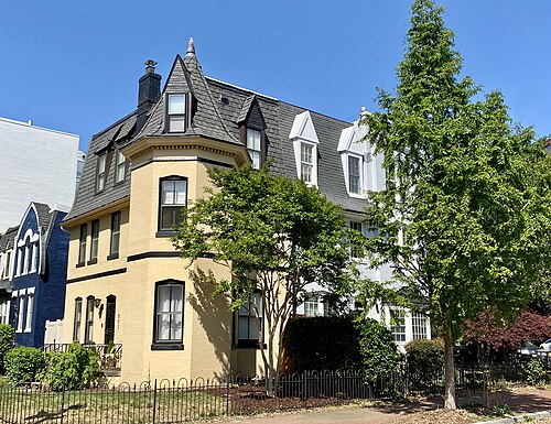 A three storey Victorian rowhouse in mustard brick with a black slate mansard roof, a turret and dormer windows, at the corner of 25th Street and New Hampshire Avenue.