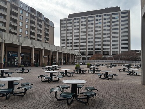 A sunken brick plaza at Columbia Plaza ringed by a shopping arcade under a low colonnade, picnic tables across the paving, an apartment tower and an office slab behind.
