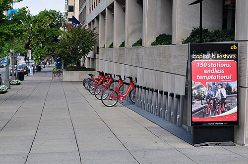A Capital Bikeshare dock lined up along a limestone clad George Washington University building, a GW banner overhead and a bike lane sign farther down the sidewalk.