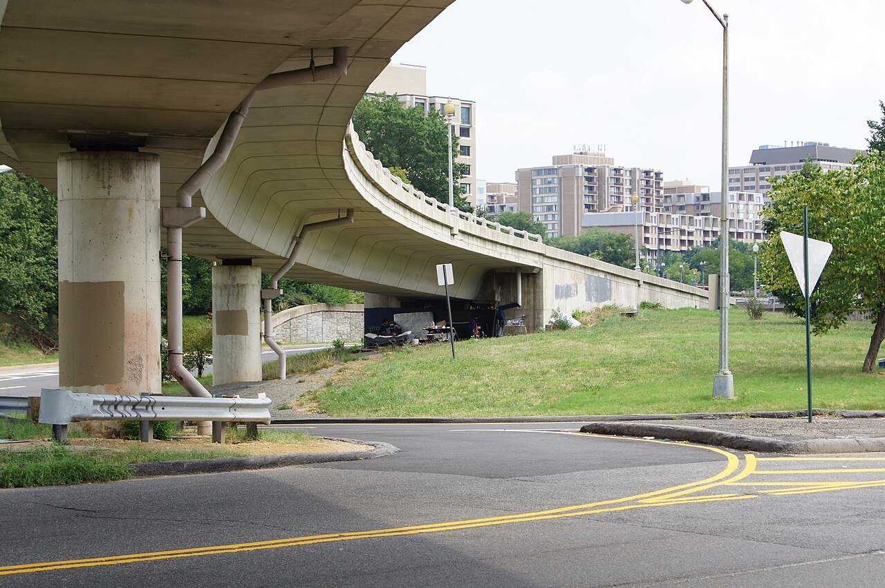 A curved elevated freeway ramp on concrete pillars near the Potomac, apartment towers across the grass beyond it, double yellow lines on the road below.