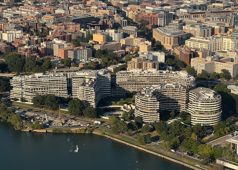 An aerial view of the Watergate complex's six curved, balconied buildings along the Potomac, courtyards and a swimming pool between the wings, downtown rooftops receding behind.
