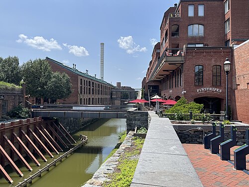 The watered canal between old brick mill buildings with a footbridge and towpath.