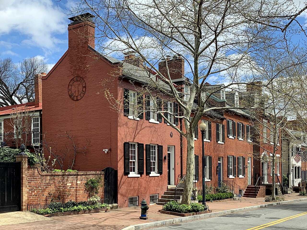 A sunlit row of Federal brick houses with black shutters, stoops, brick sidewalk and street lamp on P Street NW.