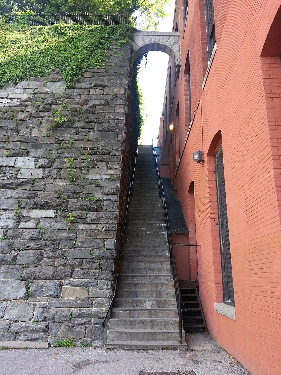 Daylight view looking up the full flight of the Exorcist steps between the stone retaining wall and the brick warehouse.