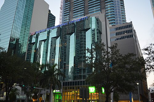 A glass office tower facade behind street trees and a street light at dusk, with FIU signage over the entrance.