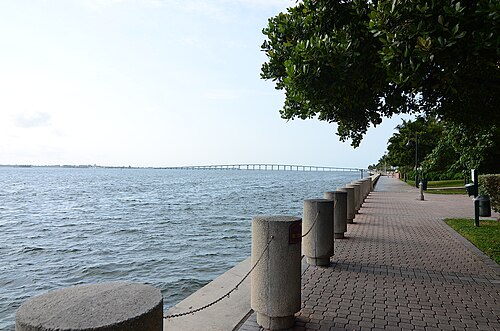 A brick paved waterfront promenade with concrete bollards along open water, a bridge on the horizon.