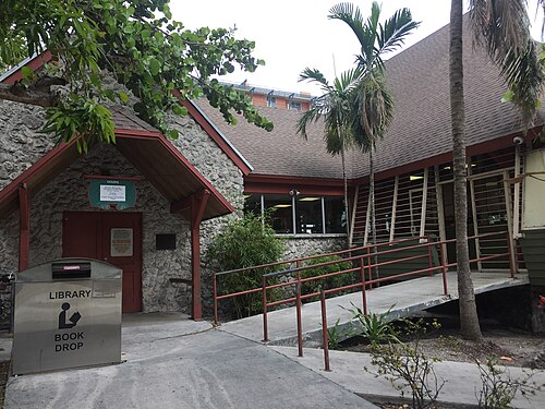 The coral rock and wood shingle entrance of the Coconut Grove Library under palm trees, with a ramp and a book drop box out front.