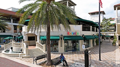 A Mediterranean Revival storefront block on Grand Avenue with green awnings, a stone fountain and a palm tree in the plaza out front.