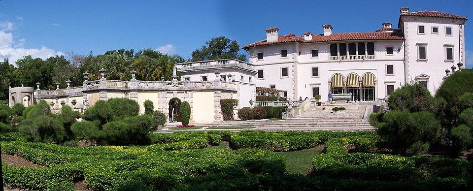 Villa Vizcaya's coral stone facade and red tile roof across a reflecting pool and clipped hedges, seen from the south garden, with a few people near the terrace steps.