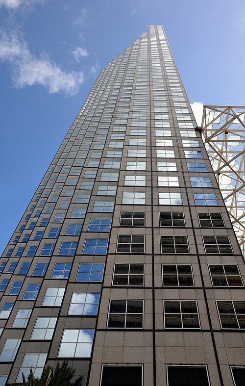 A glass curtain wall office tower seen straight up from the sidewalk, alternating bands of clear and reflective glass climbing past a construction crane.