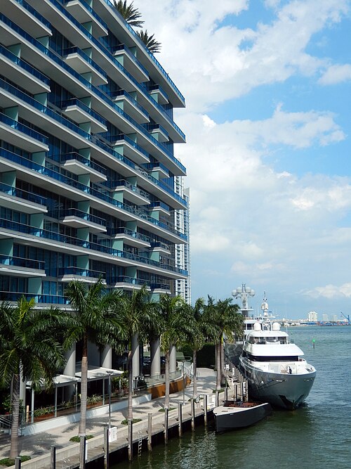A balcony fronted condominium tower along the Miami River, blue glass railings and palm trees over a paved riverwalk, a yacht moored at the dock.