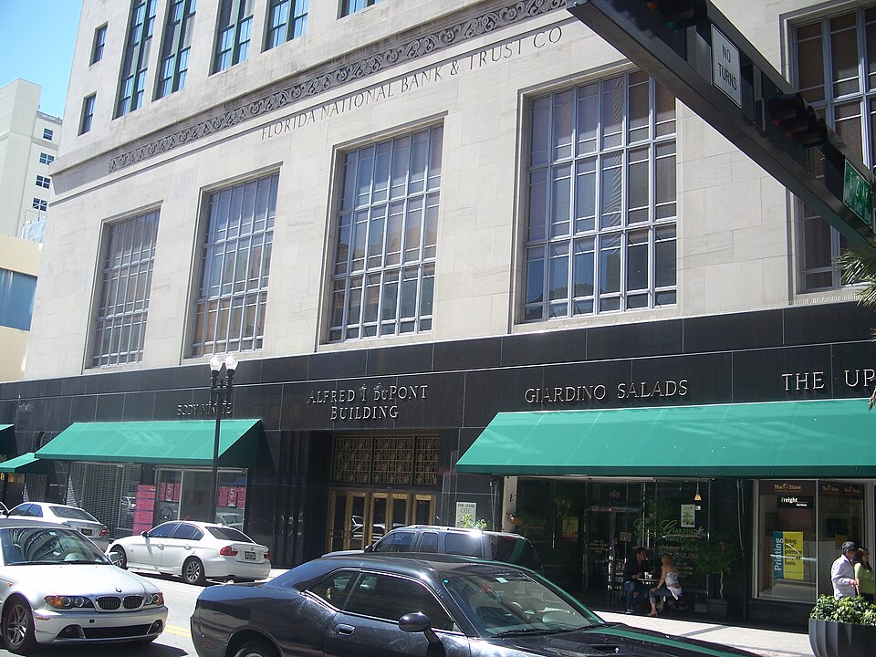 The Alfred I. DuPont Building's ground floor on East Flagler Street, ALFRED I DUPONT BUILDING carved into the black granite base under green fabric awnings, with a stone tower rising behind.