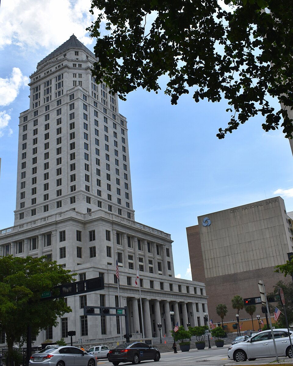 The Miami-Dade County Courthouse tower rising over West Flagler Street at NW 1st Avenue, a stepped terra cotta and stone tower with a pyramid cap over rows of narrow windows, cars and a columned base at street level.