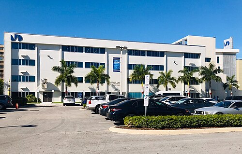The white stucco entrance of Miami Dade College's Hialeah campus, ribbon windows above a row of royal palms and a parking lot in front.
