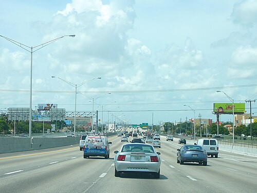 Six lanes of the Palmetto Expressway running straight beneath a row of overhead billboards and mast arm signals.