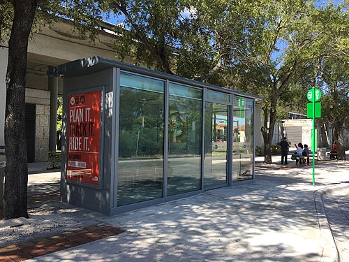 A glass sided Metrobus shelter under an elevated rail guideway, with a green bus stop pole on the sidewalk beside it.