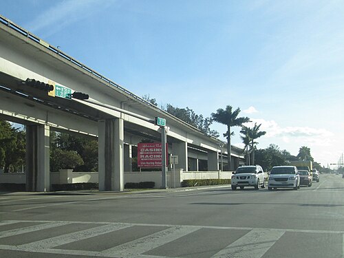 An elevated rail line on concrete piers over a wide numbered street, with a red sign at the roadside pointing to Hialeah Park Casino's entrances.