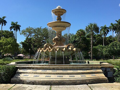 A tiered stone fountain in Hialeah Park's gardens, water running down two basins, ringed by royal palms.