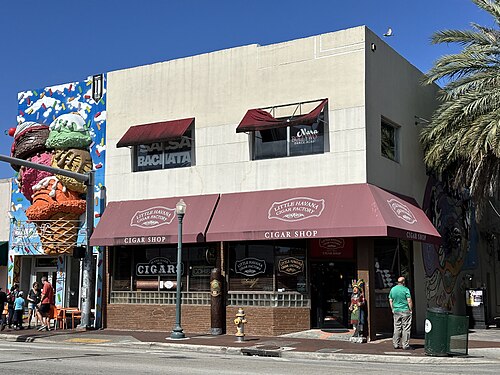 The Little Havana Cigar Factory's corner storefront, dark red awnings over glass block windows, a giant cigar column and a carved wooden figure by the door.