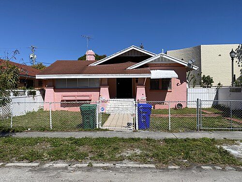 A pink one storey bungalow behind a chain link fence, a shingled porch gable and a brick chimney, a taller stucco building's blank wall behind it.