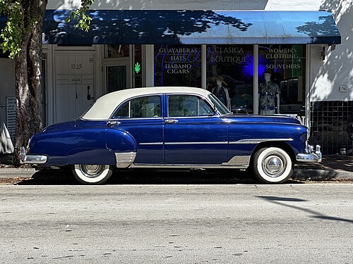 A dark blue vintage sedan parked at the curb outside a boutique window advertising guayaberas, hats and cigars, mannequins visible behind the glass.
