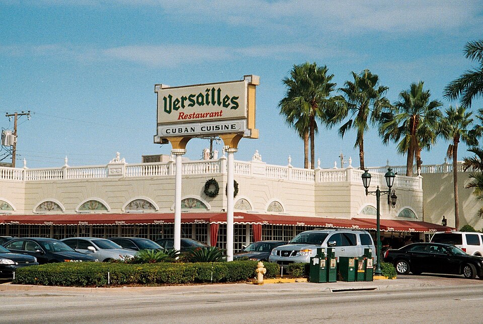Versailles restaurant's white colonnaded facade, arched window openings and lit marquee sign on Calle Ocho, cars parked along the curb under royal palms.