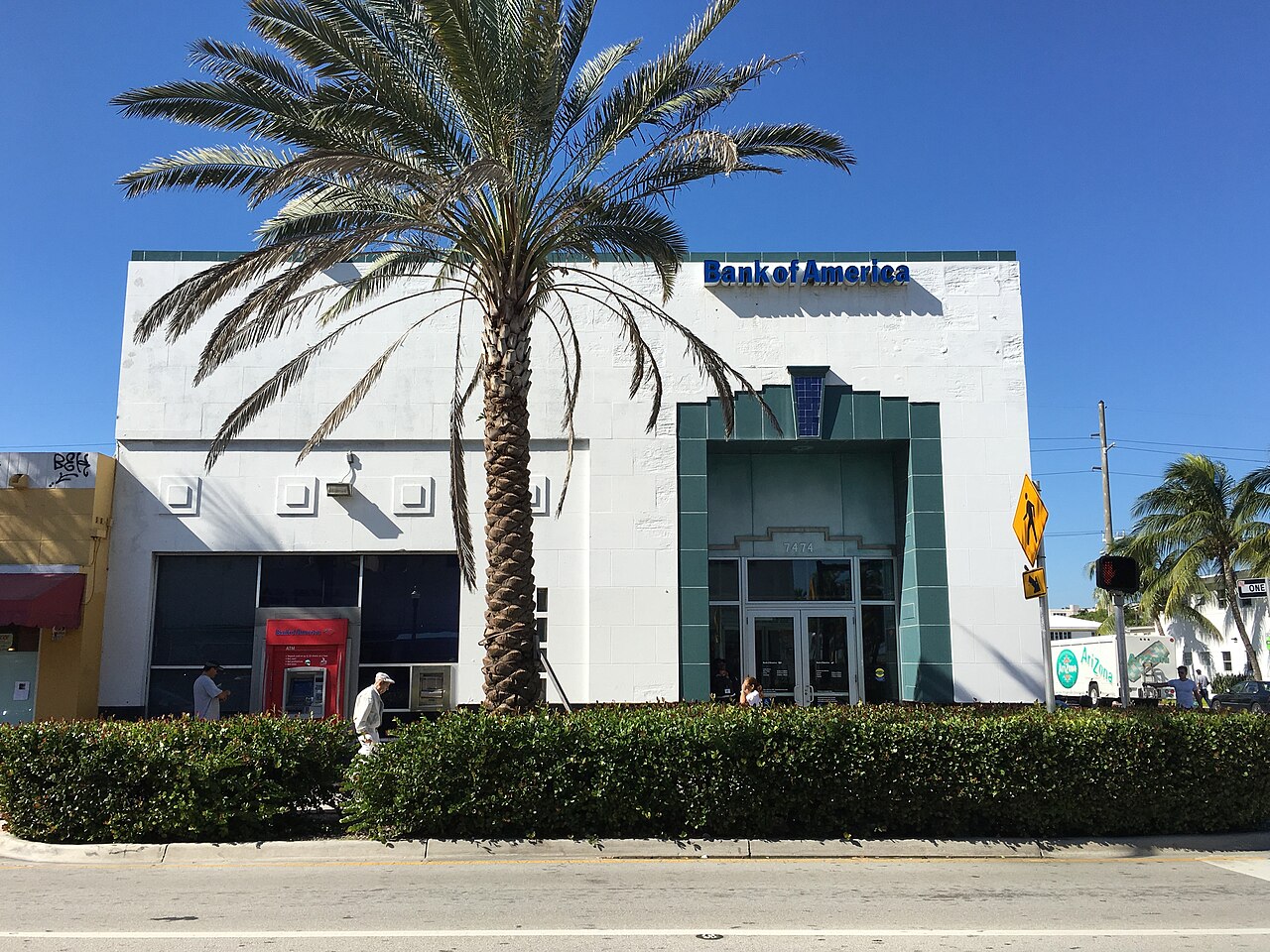 A 1950 bank building at 7474 Collins Avenue, white stucco with a teal tiled entrance surround and a Bank of America sign, an address plaque reading 7474 over the door.