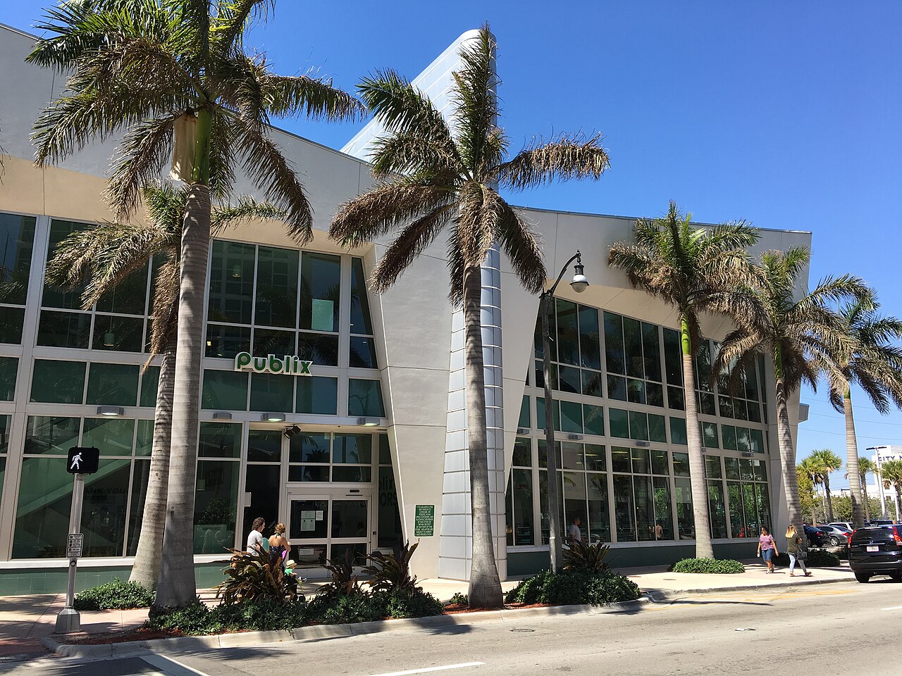 A Publix supermarket with an angular white canopy and green glazed storefront, royal palms along the sidewalk out front.