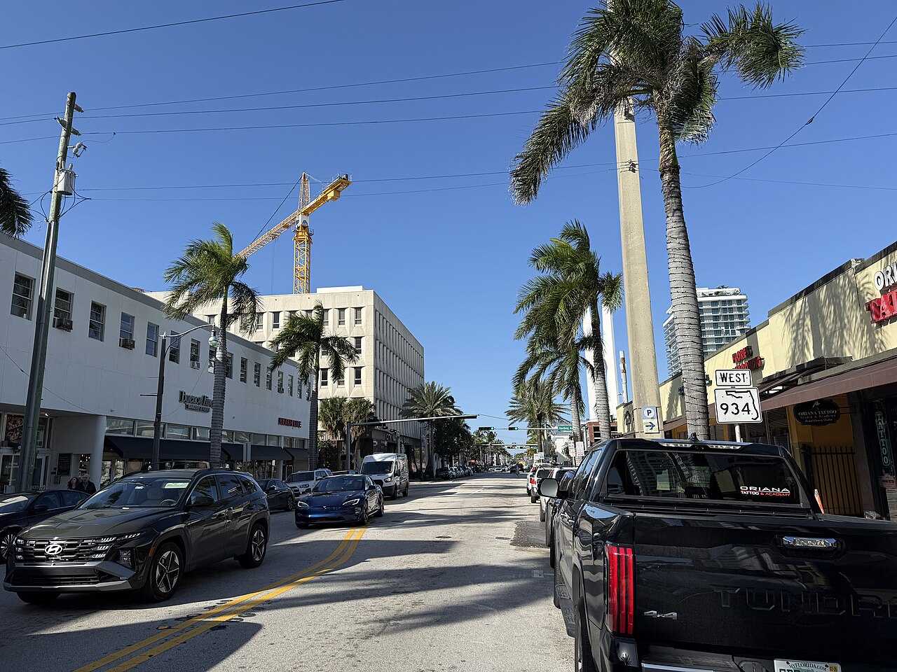 71st Street looking west from its intersection with Collins Avenue, low white commercial buildings, a construction crane behind them and palms lining the road.