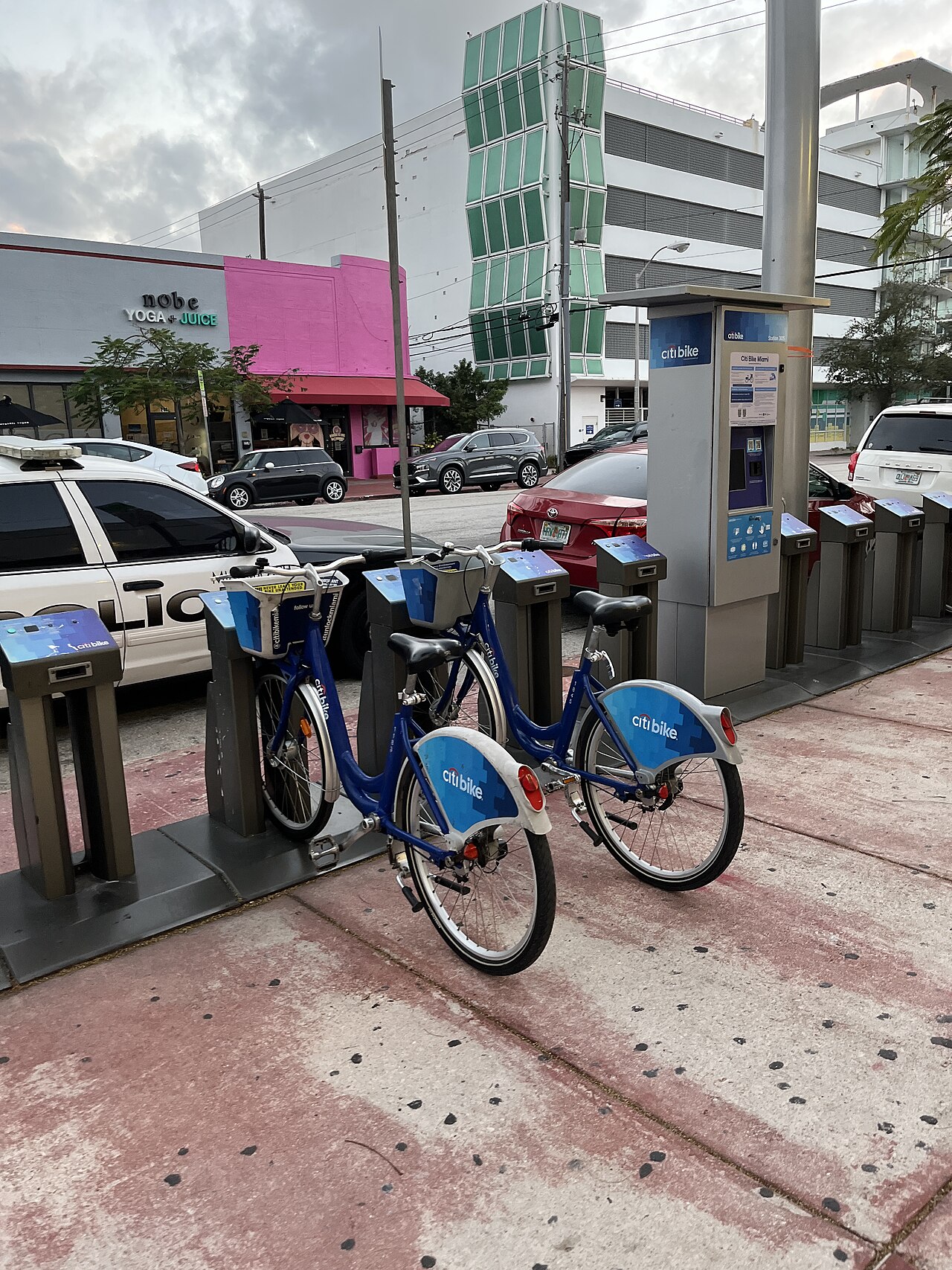 A Citi Bike station on a commercial corner, a pink stuccoed storefront and a green glazed parking garage behind, a police car at the kerb.