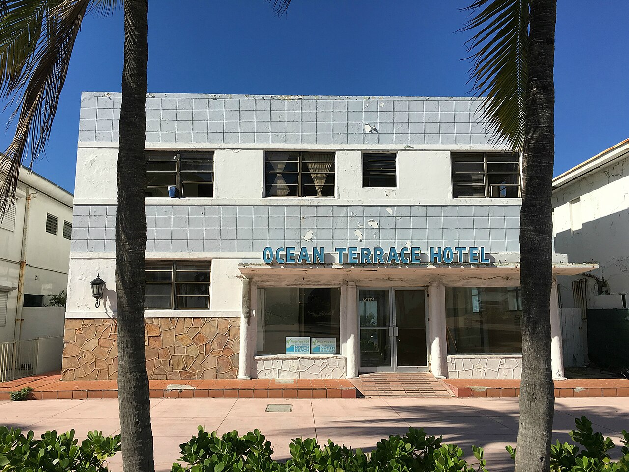 A two storey Art Deco building on Ocean Terrace, pale blue tiled facade with signage reading OCEAN TERRACE HOTEL, twin palm trees framing the entrance and a pink tiled pedestrian promenade in front.