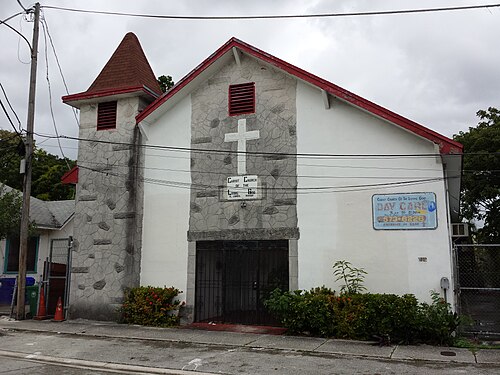 A grey fieldstone church with a conical red roofed turret and a white cross set into the gable over the entrance.