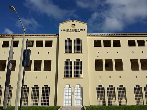 A three storey cream school building with tall grilled windows and the school's name cast into the pediment above the doors.