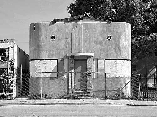 A rounded streamline moderne corner building in painted concrete, a band of glass block windows, boarded doors behind a chain link fence.