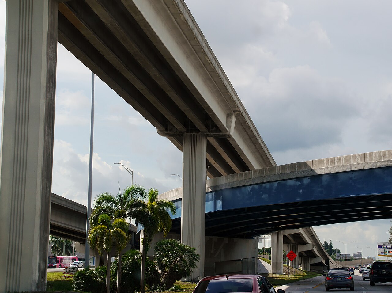 Elevated highway decks stacked on concrete columns, palm trees and cars on the roadway below.
