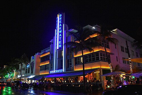 The Breakwater Hotel's vertical blue neon tower over a stepped Streamline Moderne front at night, palms lit from below and outdoor tables under umbrellas.