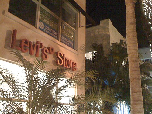 A raised copper Levi's Store sign mounted on a stucco corner at night, a leasing agent's lit window sign above it, a palm trunk close in the foreground.