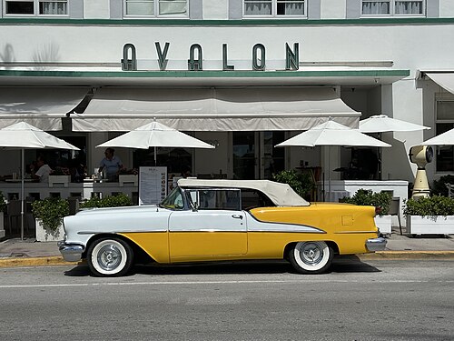 The Avalon Hotel's green striped awning and chrome AVALON lettering over a row of sidewalk umbrellas, a vintage convertible parked at the curb.