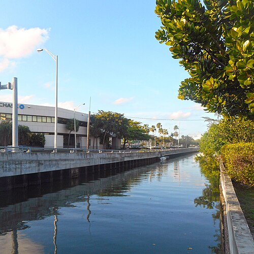 The Collins Canal looking northeast, a low modern building and street lamps along the seawall, palms and mangrove-like shrubs on the near bank.