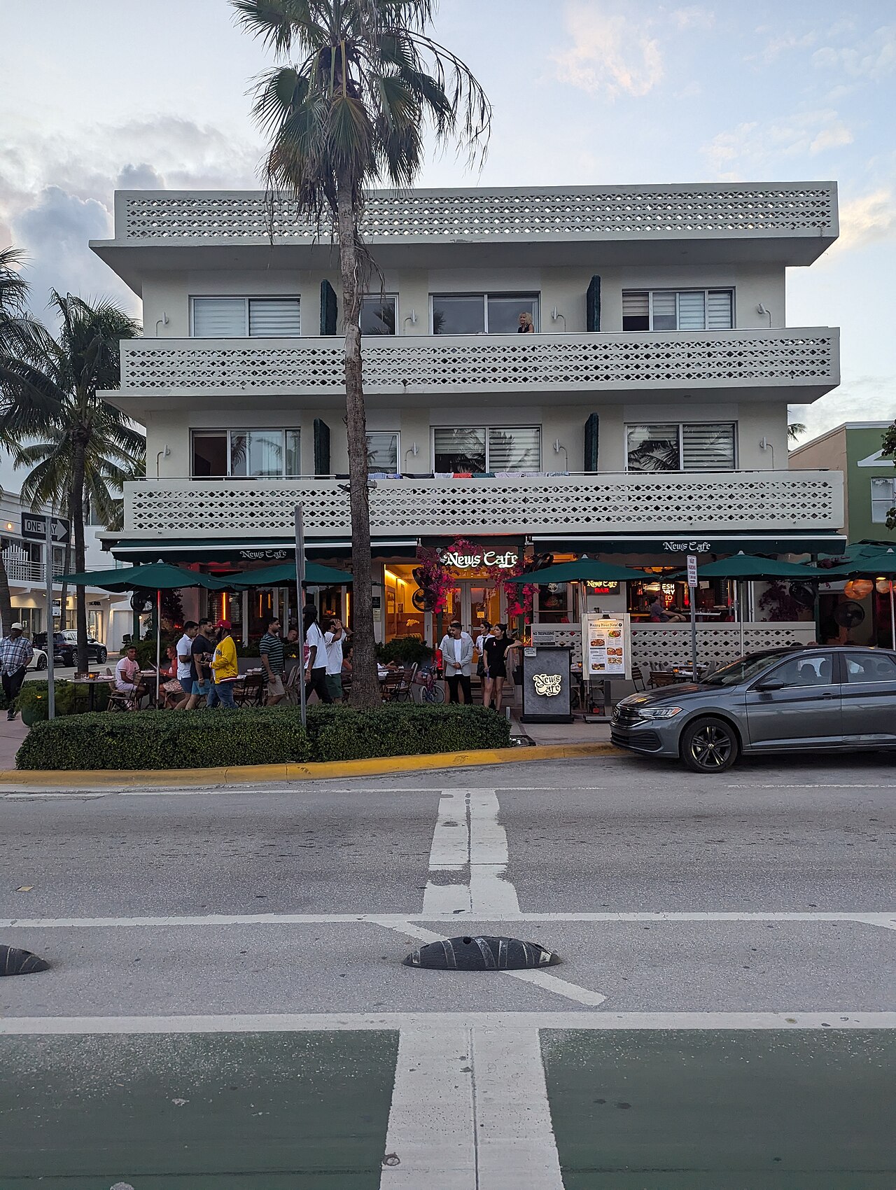 A three storey lattice balconied building on the Ocean Drive corner at Eighth Street, the News Cafe's green awnings and outdoor tables below, palms and a parked car on the street in front.