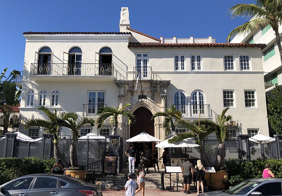 Casa Casuarina's Mediterranean Revival front, arched windows, a carved stone doorway and iron balconies behind palms and a low fence, a handful of visitors at the gate.