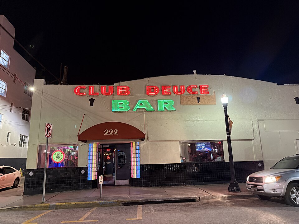 A plain white stucco corner bar under red CLUB DEUCE and green BAR neon lettering, the number 222 on a burgundy awning over the door.