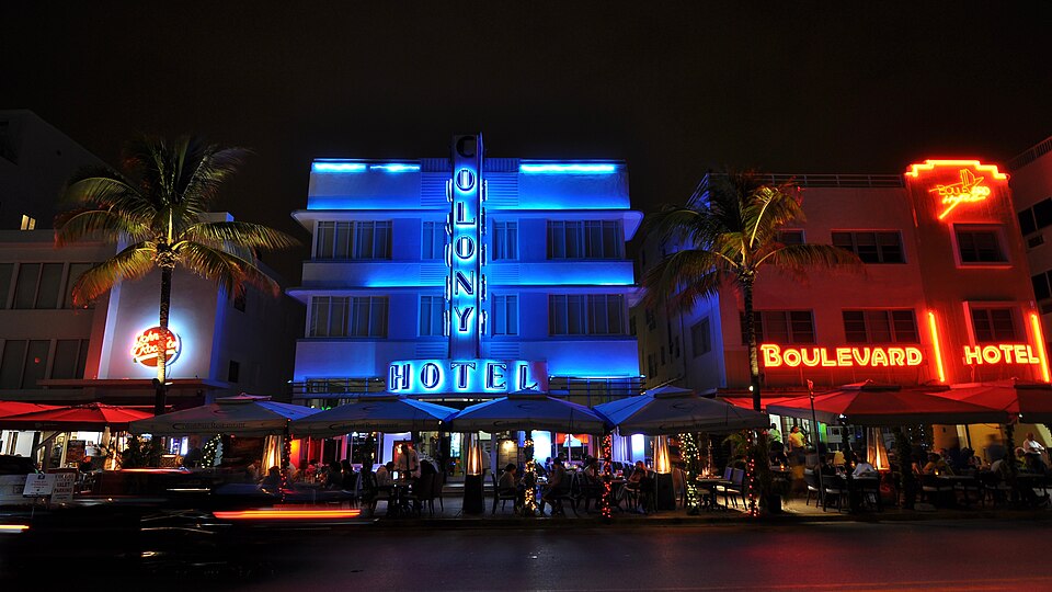 The Colony Hotel's blue vertical neon sign lit over a curved corner at night, the Boulevard Hotel's red neon next door, palms and sidewalk cafe umbrellas along the curb.